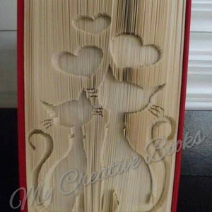 Peut inclure: Une sculpture d'art de livre représentant deux chats avec des cœurs au-dessus d'eux. Le livre est plié en un design 3D et a une couverture rouge. Le texte "My Creative Books" est visible sur le côté droit du livre.
