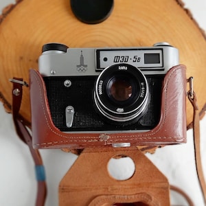 Puede incluir: Una cámara vintage plateada y negra con una funda de cuero marrón y una correa. La cámara tiene un objetivo con una tapa negra y una pequeña pantalla digital que muestra "0.30 - 5.8".