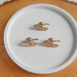May include: Three gold-colored tank earrings on a white surface. The tanks are detailed with a raised design.