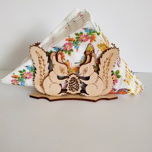 May include: Wooden napkin holder featuring two squirrels facing each other, holding a pine cone. The squirrels are carved in a detailed style with visible fur and whiskers. The holder is designed to hold a stack of napkins.