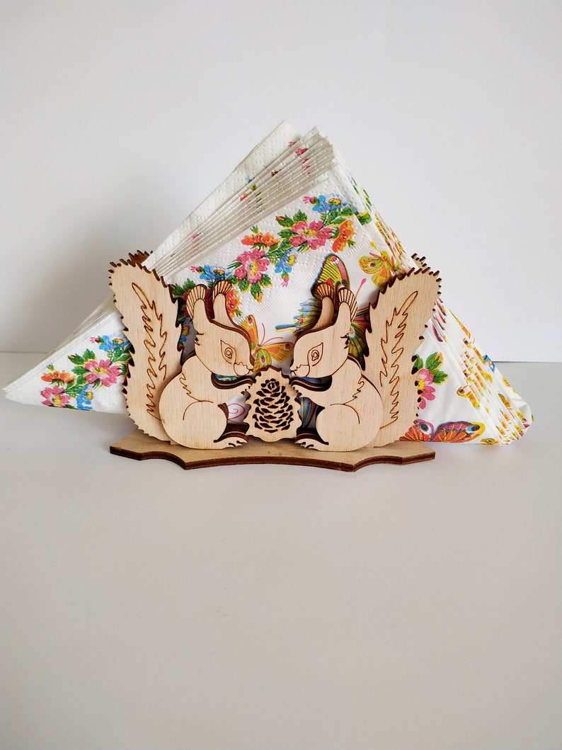 May include: Wooden napkin holder featuring two squirrels facing each other, holding a pine cone. The squirrels are carved in a detailed style with visible fur and whiskers. The holder is designed to hold a stack of napkins.