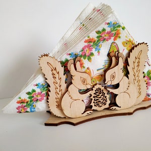 May include: Wooden napkin holder featuring two squirrels holding a pine cone. The squirrels are carved in a light brown wood and have detailed features. The holder is designed to hold a stack of napkins.