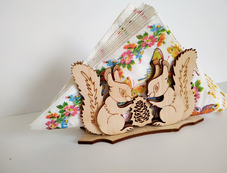 May include: Wooden napkin holder featuring two squirrels holding a pine cone. The squirrels are carved in a light brown wood and have detailed features. The holder is designed to hold a stack of napkins.