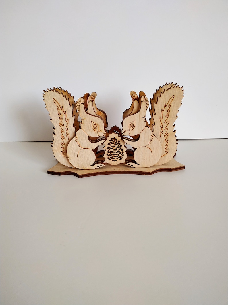 May include: Wooden napkin holder featuring two squirrels facing each other, holding a pine cone. The squirrels are carved in a light brown wood and have detailed features.