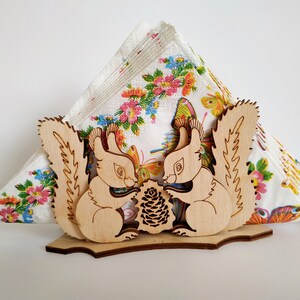 May include: Wooden napkin holder featuring two squirrels holding a pine cone. The squirrels are carved in a light wood and have detailed features. The holder is decorated with a floral pattern.