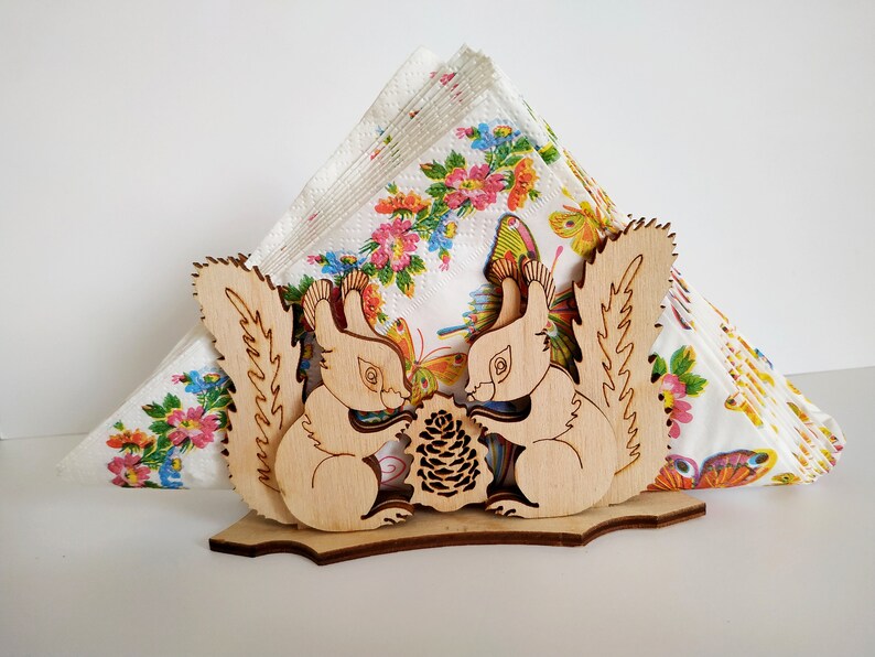 May include: Wooden napkin holder featuring two squirrels holding a pine cone. The squirrels are carved in a light wood and have detailed features. The holder is decorated with a floral pattern.
