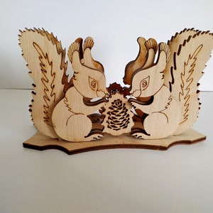May include: Wooden napkin holder featuring two squirrels facing each other, holding a pine cone. The squirrels are carved in a detailed, whimsical style.
