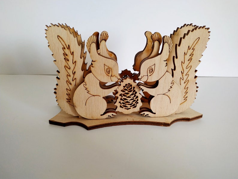 May include: Wooden napkin holder featuring two squirrels facing each other, holding a pine cone. The squirrels are carved in a detailed, whimsical style.