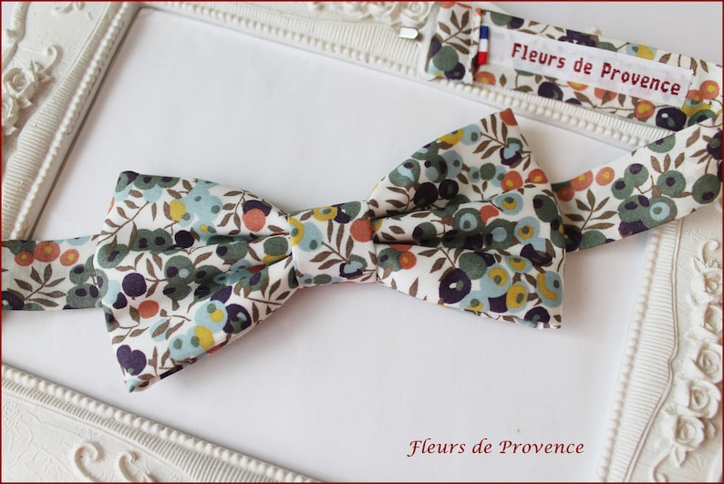 Puede incluir: Una pajarita blanca con un patr&oacute;n floral en tonos de verde, azul, naranja y marr&oacute;n. La pajarita est&aacute; hecha de tela y tiene una etiqueta "Fleurs de Provence" adjunta.