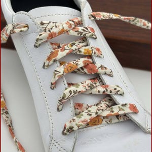 May include: White trainers with floral patterned shoelaces. The laces are a cream background with orange and pink flowers. The text "Fleurs de Provence" is visible at the bottom of the image.