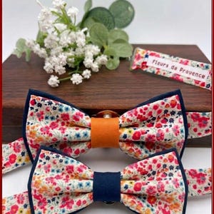 May include: Two floral bow ties with navy blue trim and straps. The fabric features a pattern of red, pink, yellow, and blue flowers on a cream background. One bow tie has an orange center band, the other has a navy blue center band. The text "Fleurs de Provence" is visible.