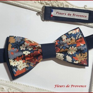 May include: A navy blue bow tie featuring a floral design in shades of orange, white, and blue. The bow tie has a dark blue adjustable strap. The text "Fleurs de Provence" is visible on the label.