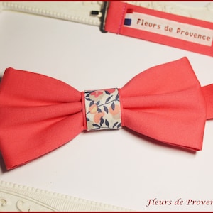 May include: A coral pink bow tie with a white and floral patterned center. The bow tie is made of fabric and has a simple design. The text "Fleurs de Provence" is printed on the bow tie.