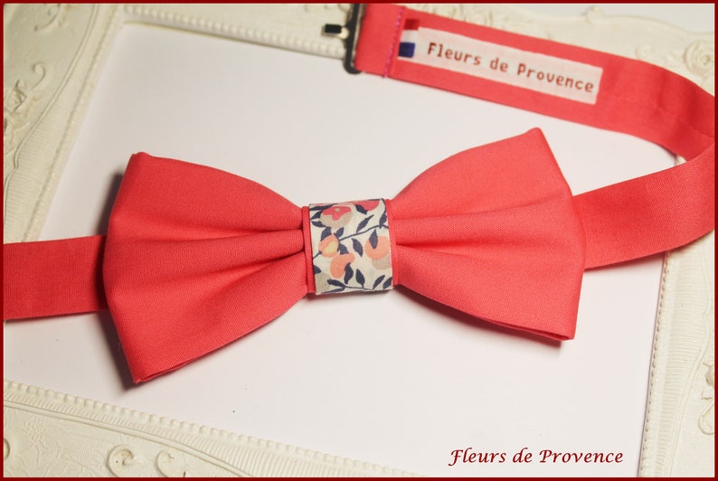 May include: A coral pink bow tie with a white and floral patterned center. The bow tie is made of fabric and has a simple design. The text "Fleurs de Provence" is printed on the bow tie.