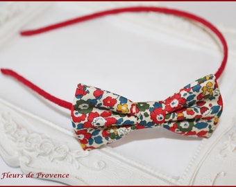May include: A red headband with a floral print bow. The bow is made of fabric with red, blue, yellow, and white flowers on a cream background.
