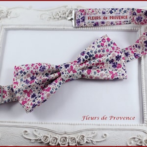 Può includere: Un papillon bianco con un motivo floreale rosa e viola. Il papillon è realizzato in tessuto e ha un cinturino regolabile abbinato con il testo "FLEURS de PROVENCE" stampato su di esso.
