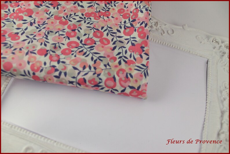 May include: A white fabric with a pink and blue floral pattern. The fabric is folded and placed on top of a white picture frame. The frame has a decorative border and the text "Fleurs de Provence" is printed on the bottom right corner.