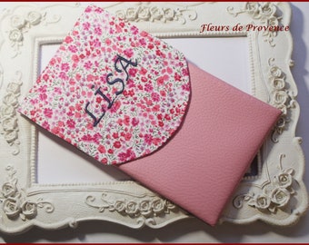 May include: A pink fabric phone case with a floral pattern and the name "Lisa" embroidered on it. The case is folded in half and is sitting on a white ornate frame.