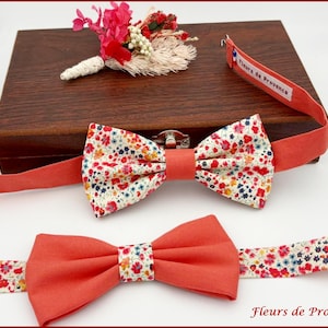 May include: Two bow ties with floral patterns and coral-colored bands. One bow tie is tied, and the other is untied. A small floral boutonniere and a wooden box are in the background. The text "Fleurs de Provence" is visible.