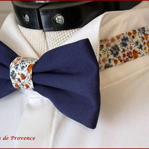 May include: A navy blue bow tie with a white and orange floral pattern. The bow tie is tied around a white shirt collar.