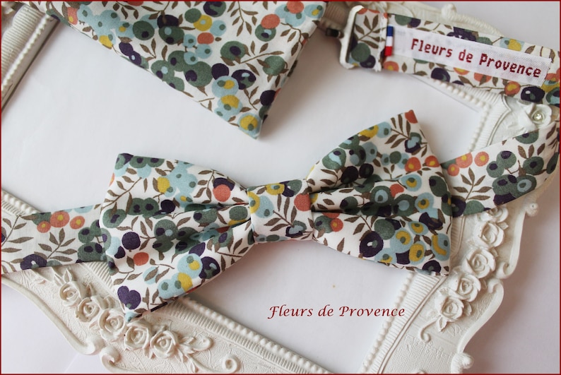 Puede incluir: Una pajarita blanca con un patr&oacute;n floral en tonos verdes, azules, naranjas y amarillos. La pajarita est&aacute; hecha de tela y tiene una etiqueta que dice "Fleurs de Provence".