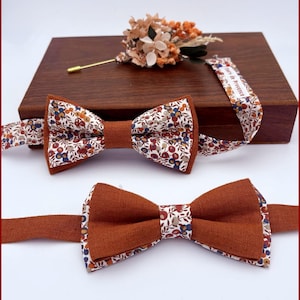 Può includere: Due papillon, uno con un motivo floreale e uno di colore ruggine pieno. I papillon sono esposti su una scatola di legno con un nastro floreale e un piccolo bouquet di fiori secchi.