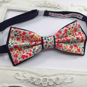 May include: A navy blue bow tie with a white background and a floral pattern in shades of red, orange, yellow, and green. The bow tie has a label that reads "FLEURS DE PROVENCE".