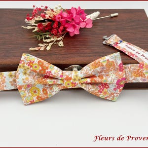 Peut inclure: Un nœud papillon à motifs floraux avec des fleurs orange, jaunes et roses. Une sangle florale assortie et une boutonnière de fleurs roses avec une épingle sont également présentes. Le nœud papillon et la boutonnière sont sur une surface en bois foncé.