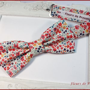 May include: A white bow tie with a floral pattern in shades of red, blue, and yellow. The bow tie is made of fabric and has a small label that says "Fleurs de Provence".