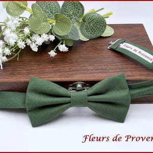 Pode incluir: Uma gravata borboleta verde com o texto "Fleurs de Provence" na parte inferior. A gravata borboleta está em uma caixa de madeira com flores brancas e folhas verdes. A alça da gravata borboleta tem o texto "Fleurs de Provence".