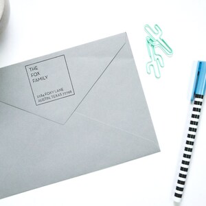 Peut inclure: Une enveloppe grise avec une étiquette d'adresse de retour qui indique "The Fox Family, Harmony Lane, Austin, Texas 78718". L'enveloppe est sur une surface blanche avec un bougeoir en béton, un stylo rayé bleu et noir et deux trombones en forme de cactus verts.
