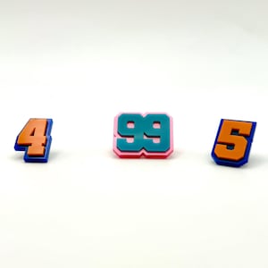 Może przedstawiać: Trzy plastikowe szpilki z numerami w kolorze pomarańczowym, turkusowym i różowym. Numery to 4, 99 i 5.