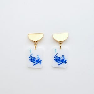 May include: A pair of gold-toned earrings. Each earring features a gold half-circle top, connected to a white rectangular charm. The charm displays a blue peacock design and the number "1".