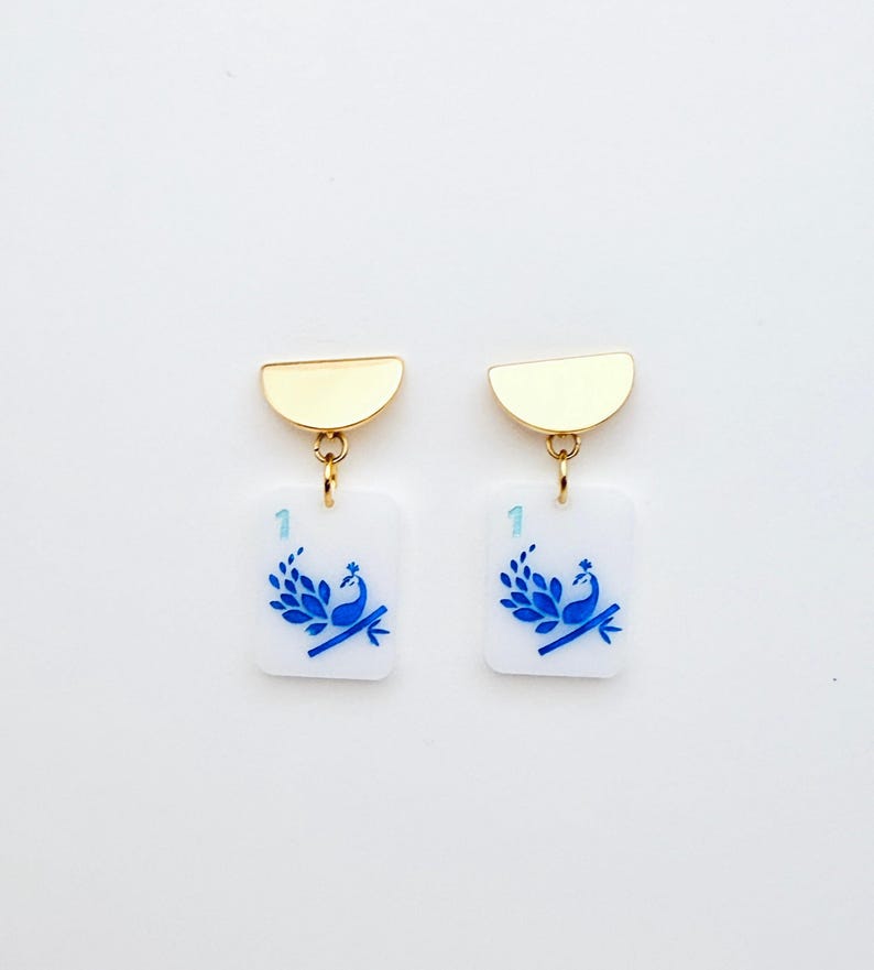 May include: A pair of gold-toned earrings. Each earring features a gold half-circle top, connected to a white rectangular charm. The charm displays a blue peacock design and the number "1".