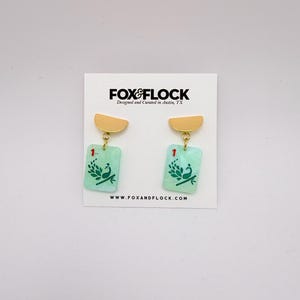 May include: A pair of earrings with a gold-colored half-circle top and a rectangular, light green bottom. The bottom of the earrings have a peacock design and the number 1. The earrings are hanging on a white card with the text "FOX & FLOCK" and "www.foxandflock.com".