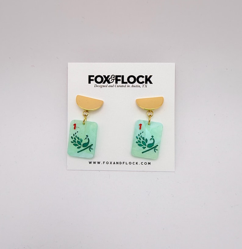 May include: A pair of earrings with a gold-colored half-circle top and a rectangular, light green bottom. The bottom of the earrings have a peacock design and the number 1. The earrings are hanging on a white card with the text "FOX & FLOCK" and "www.foxandflock.com".
