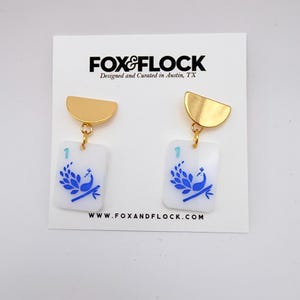 May include: A pair of gold-toned, half-circle earrings with rectangular white pendants. Each pendant features a blue peacock design and the number "1". The earrings are from Fox & Flock, designed and curated in Austin, TX.