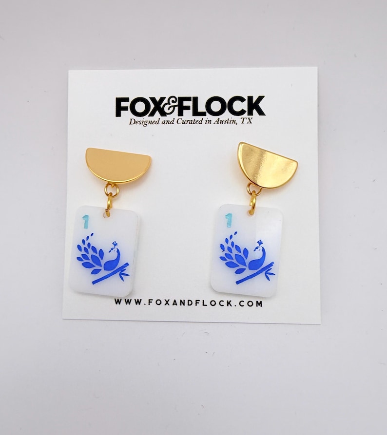 May include: A pair of gold-toned, half-circle earrings with rectangular white pendants. Each pendant features a blue peacock design and the number "1". The earrings are from Fox & Flock, designed and curated in Austin, TX.