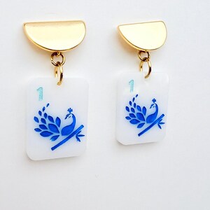 May include: A pair of gold-tone earrings. Each earring features a gold half-circle top connected to a white rectangular charm. The charm has a blue peacock design and the number "1".