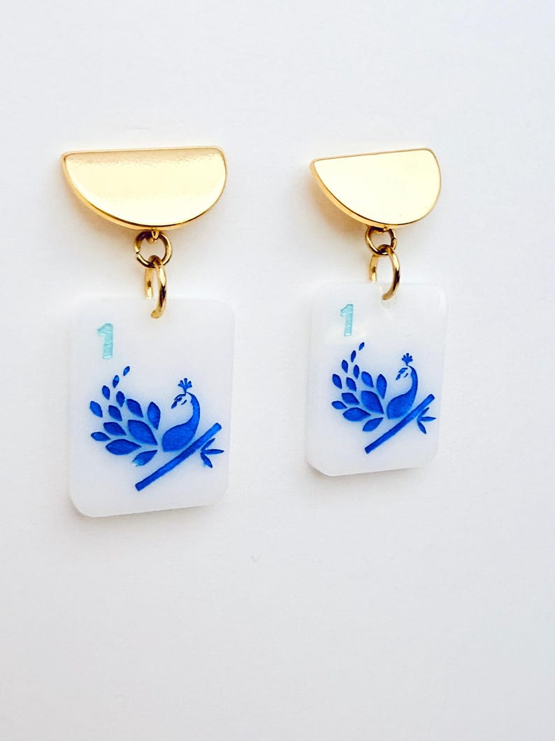 May include: A pair of gold-tone earrings. Each earring features a gold half-circle top connected to a white rectangular charm. The charm has a blue peacock design and the number "1".