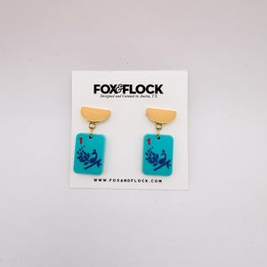 May include: A pair of turquoise earrings with gold-colored accents. Each earring features a rectangular pendant with a peacock design and the number "1". The earrings are displayed on a white card with the "FOX&FLOCK" logo.