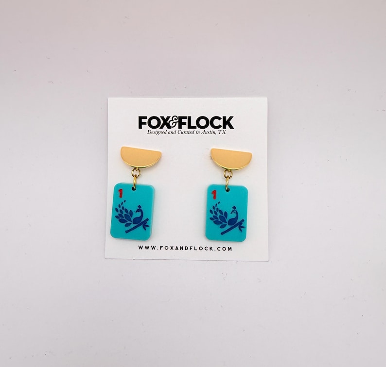 May include: A pair of turquoise earrings with gold-colored accents. Each earring features a rectangular pendant with a peacock design and the number "1". The earrings are displayed on a white card with the "FOX&FLOCK" logo.