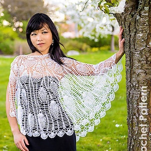 Peut inclure: Un châle en crochet blanc avec un motif de plume de paon. Le châle est porté par une personne debout dans un jardin avec un arbre en arrière-plan.