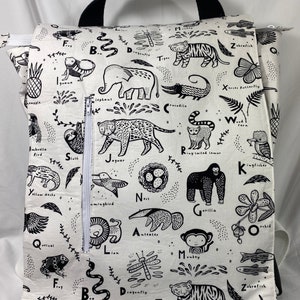 May include: Black and white backpack with a zippered pocket. The backpack features a repeating pattern of animals and letters of the alphabet. The animals include a tiger, a jaguar, a gorilla, a monkey, a sloth, a hummingbird, a dragonfly, a kingfisher, a zebra fish, a butterfly, a crocodile, an anteater, a lion, a frog, a gecko, a quetzal, a pineapple, an iguana, an elephant, a  lemur, a bird, a nest, and a snake.
