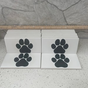 May include: White ceramic tiles with a black paw print design. The tiles are arranged in stacks, showcasing the paw print graphic. The paw prints are a dark gray color, contrasting against the white background. The tiles are likely for home decor or craft projects.