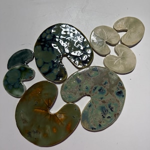 May include: A collection of ceramic lily pads in various sizes and colors. The pads range from deep green and teal to muted olive and cream, with unique speckled and mottled patterns. The glossy finish enhances the naturalistic design.