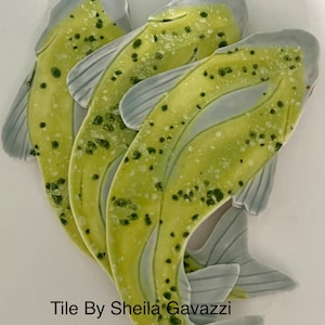May include: Three ceramic fish tiles in shades of green and blue with a speckled glaze. The tiles are arranged in a stack, with the tails of the fish overlapping. The text "Tile By Sheila Gavazzi" is visible at the bottom of the image.