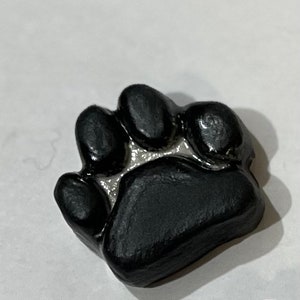 May include: A black paw print charm with a silver outline. The charm is made of clay and has a textured surface.