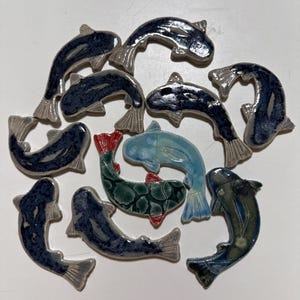May include: A collection of eleven ceramic fish-shaped tiles arranged in a circular pattern. The tiles feature various glaze colors including deep navy blue, mottled green with red accents, and light blue, each with a glossy finish and defined fins.