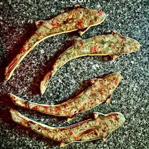 May include: Four ceramic fish tiles with a speckled pattern in shades of green, red, and gold. The tiles are arranged on a textured, dark gray surface. The text "Tiles By Sheila Gavazzi & Son" is at the top.